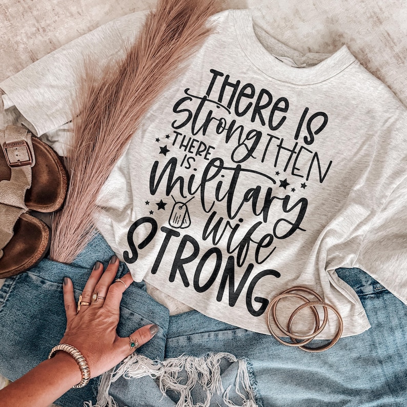 May include: A light gray short-sleeved t-shirt with black text that reads There is strong then there is military wife strong, featuring a small dog tag graphic, paired with blue denim shorts, brown sandals, and three thin gold-toned bracelets.