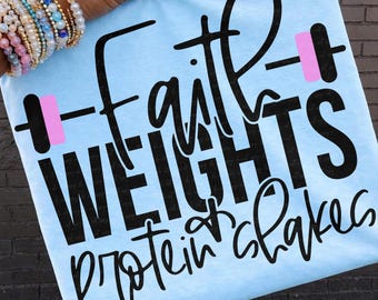 Puede incluir: Camiseta azul claro con la frase "Faith Weights Protein Shakes" en negro. La palabra "Faith" está estilizada con un gráfico de barra rosa. Prenda de vestir informal.