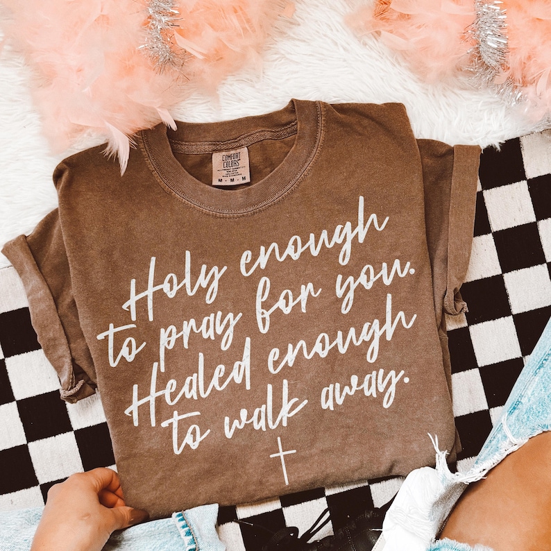 May include: A brown Comfort Colors t-shirt with white text that reads "Holy enough to pray for you. Healed enough to walk away." A small white cross is below the text. The shirt is on a black and white checkered surface.
