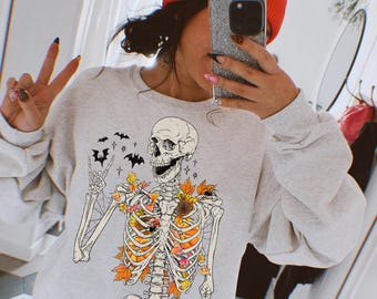 Könnte beinhalten: Ein graues Sweatshirt mit einem Skelett, das Herbstblätter trägt und ein Peace-Zeichen macht. Das Skelett lächelt und hat einen Schädel mit einer schwarzen Augenhöhle. Der Text "Elena Maria Designs" und "Original Designer" befindet sich am unteren Rand des Bildes.