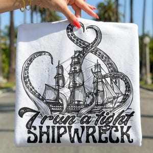 Könnte beinhalten: Weißes Sweatshirt mit einer Schwarz-Weiß-Illustration eines Schiffes, das von einem Oktopus angegriffen wird. Der Text "I run a tight SHIPWRECK" ist darunter gedruckt. Das Sweatshirt wird von einer Person gehalten.