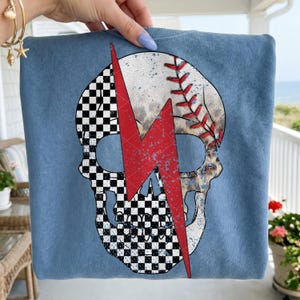 Peut inclure: T-shirt bleu clair avec un motif de crâne. Le crâne est divisé par un éclair rouge. Une moitié du crâne est un ballon de baseball, l'autre moitié est un motif à carreaux noir et blanc.