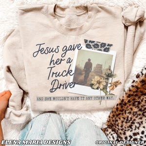 Può includere: Felpa beige con il testo "Jesus gave her a Truck Driver" e una foto di un uomo che si allontana da un camion. Il testo "And she wouldn't have it any other way." è sotto la foto.