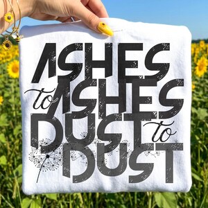 May include: White t-shirt with the phrase "Ashes to Ashes, Dust to Dust" in large, dark gray block letters. The design includes a dandelion graphic. The shirt is held up against a background of sunflowers.