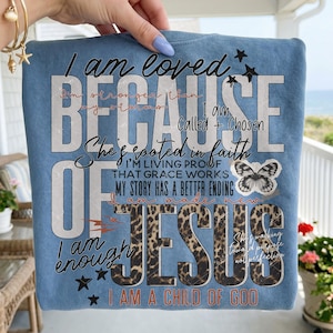 May include: Light blue t-shirt with the text "I am loved BECAUSE OF JESUS I am a child of God". The design includes a butterfly, stars, and leopard print accents. The shirt has a crew neck.