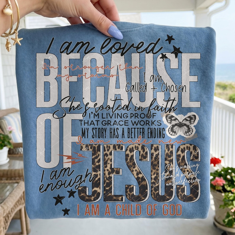 May include: Light blue t-shirt with the text "I am loved BECAUSE OF JESUS I am a child of God". The design includes a butterfly, stars, and leopard print accents. The shirt has a crew neck.