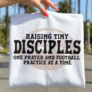 May include: White t-shirt with the text "RAISING TINY DISCIPLES ONE PRAYER AND FOOTBALL PRACTICE AT A TIME." The text is in black, with a football graphic in the background.