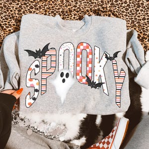 May include: A heather gray crewneck sweatshirt featuring the word SPOOKY in large, patterned letters. The design includes black bats, a white ghost, and various patterns like orange and pink stripes, checkered squares, and small pumpkins.