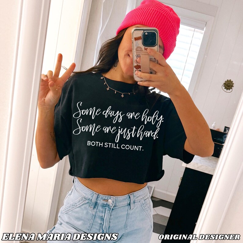 May include: Black cropped t-shirt with the text "Some days are holy. Some are just hard. BOTH STILL COUNT." in white script. The model is wearing a pink beanie and light blue jeans. The text "ELENA MARIA DESIGNS" is at the bottom.