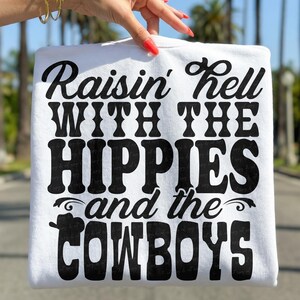 May include: A white sweatshirt with the text "Raisin' Hell WITH THE HIPPIES and the COWBOYS" in bold black lettering. The sweatshirt is held up by a hand with red nail polish, against a blurred outdoor background.