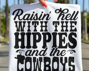 Op de afbeelding: Een witte sweatshirt met de tekst "Raisin' Hell WITH THE HIPPIES and the COWBOYS" in dikke, zwarte letters. De sweatshirt wordt vastgehouden door een hand met rode nagellak, tegen een vage buitenachtergrond.