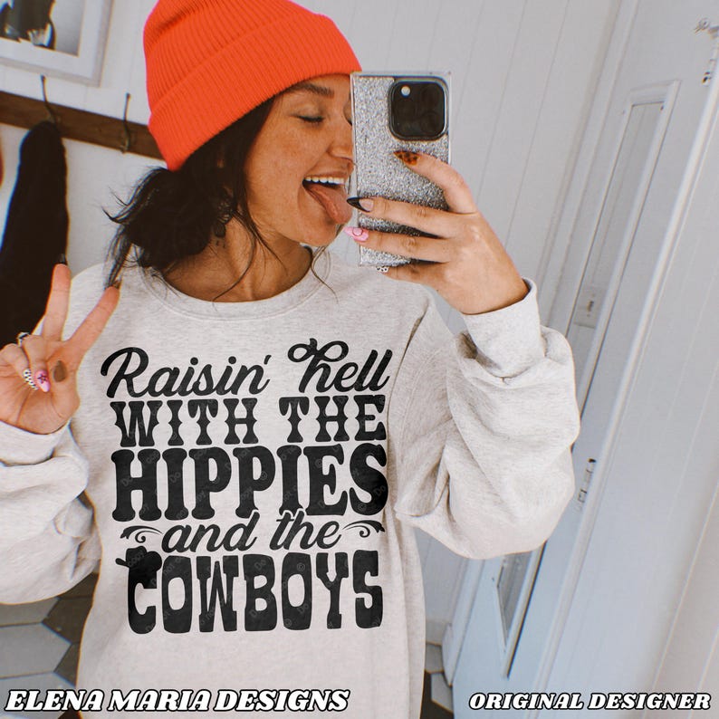 May include: A light gray sweatshirt with the text "Raisin' Hell With The Hippies and the Cowboys" in black. The sweatshirt is paired with an orange beanie. The words "ELENA MARIA DESIGNS" and "ORIGINAL DESIGNER" are at the bottom.