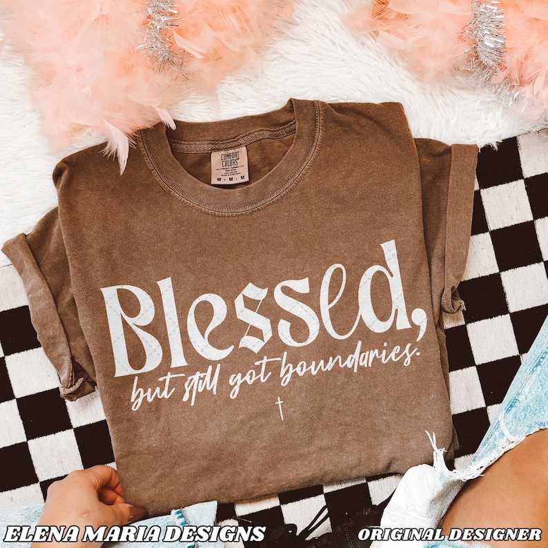 May include: A brown t-shirt with the text "Blessed, but still got boundaries." in white lettering. The shirt has a distressed look and a small cross symbol below the text.