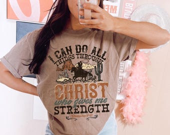 May include: A brown t-shirt with a vintage-style graphic design featuring a cowboy riding a horse. The graphic includes the text "I can do all things through Christ who gives me strength" and a scripture reference.