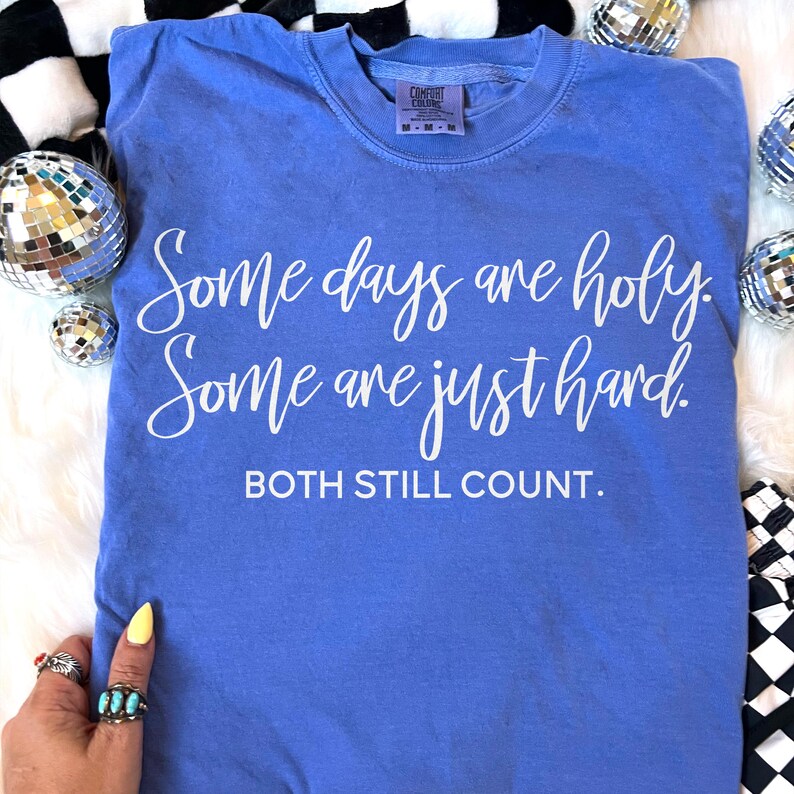 May include: A blue Comfort Colors t-shirt with white text that reads "Some days are holy. Some are just hard. Both still count." The shirt has a crew neck and a tag at the neckline. The shirt is surrounded by disco balls and a black and white checkered pattern.
