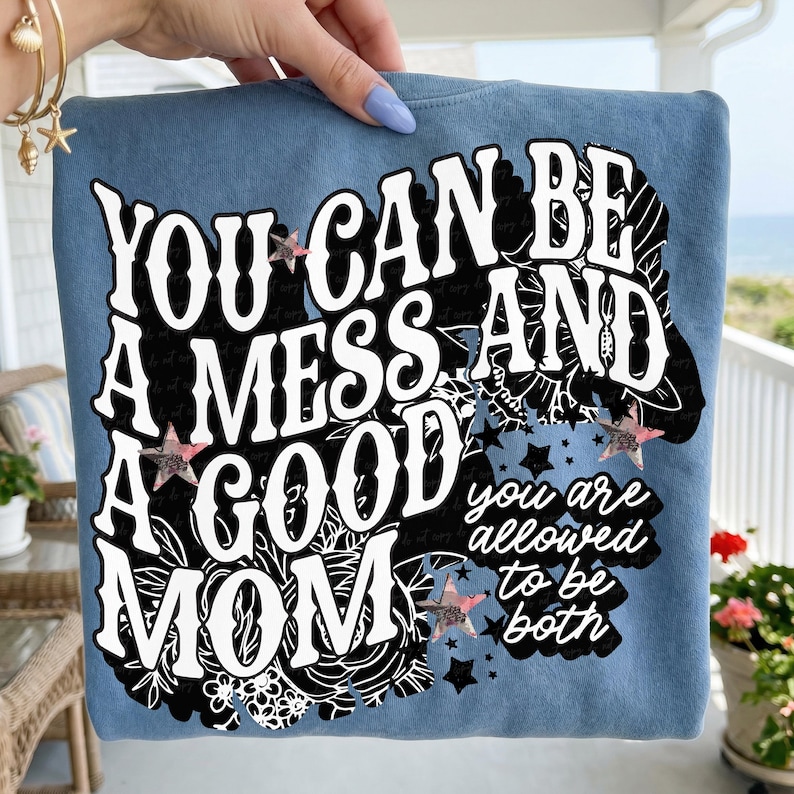 May include: A blue sweatshirt with the text "YOU CAN BE A MESS AND A GOOD MOM" in white, with black floral accents and star details. The text "you are allowed to be both" is also included.