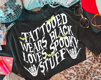 May include: A black t-shirt featuring white text that reads TATTOOED WEARS BLACK LOVES SPOOKY STUFF with small green bat graphics and two white skeleton hand illustrations. The shirt is surrounded by a disco ball, white sneakers, sunglasses, and a leopard print fabric.