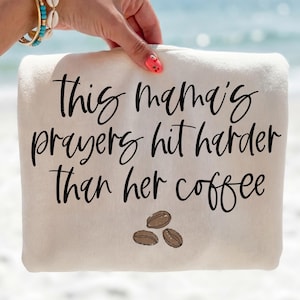 May include: A cream-colored towel with the text "This mama's prayers hit harder than her coffee" in black script. Three brown coffee beans are at the bottom. The towel is held by a hand with a turquoise bracelet.