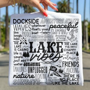 Peut inclure: T-shirt blanc avec texte noir, avec des phrases comme "Lake Vibes", "Sunshine" et "Unplugged". Le motif comprend des illustrations d'arbres, d'une ancre et d'une pagaie. Le t-shirt est tenu par une main.