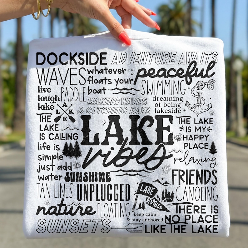 Peut inclure: T-shirt blanc avec texte noir, avec des phrases comme "Lake Vibes", "Sunshine" et "Unplugged". Le motif comprend des illustrations d'arbres, d'une ancre et d'une pagaie. Le t-shirt est tenu par une main.