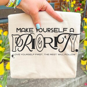 May include: Cream-colored tote bag with the text "MAKE YOURSELF A PRIORITY." in black, stylized font. Below the main text is the phrase "LOVE YOURSELF FIRST, THE REST WILL FOLLOW." The bag is held by a hand with light blue nail polish.