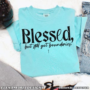 May include: A light blue t-shirt with the text "Blessed, but still got boundaries." in black lettering. A small cross is below the text.