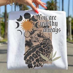 May include: White sweatshirt featuring a graphic design with a leopard, sun, moon phases, and the text "You are worthy always." The design incorporates shades of black, brown, and orange. The sweatshirt is held up by a person.