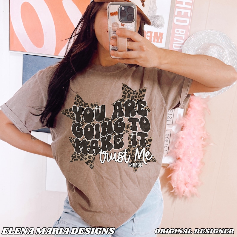 May include: A brown t-shirt with the text "YOU ARE GOING TO MAKE IT trust Me" in a black and white design. The text is set against a leopard print starburst background. The words "ELENA MARIA DESIGNS" and "ORIGINAL DESIGNER" are at the bottom.