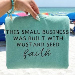 May include: A seafoam green sweatshirt with the text "THIS SMALL BUSINESS WAS BUILT WITH MUSTARD SEED faith" in black lettering. The sweatshirt is held up against a beach background with blue umbrellas and the ocean.