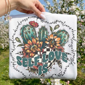 May include: White sweatshirt featuring a colorful graphic of cacti, flowers, and a checkered heart. The words "SELF LOVE IS" are in teal letters. The design is surrounded by words like "kindness" and "strength".