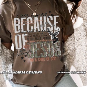 May include: A brown t-shirt with the words "I am loved because of Jesus" in white and camouflage lettering. The shirt features a deer graphic and additional text, including "I am a child of God."