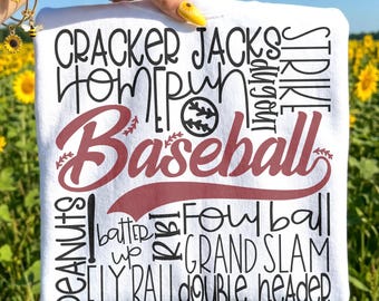 Op de afbeelding: Wit T-shirt met een honkbalthema. Het shirt toont het woord "Baseball" in een bordeauxrode letter, omringd door honkbalgerelateerde woorden zoals "Home Run" en "Grand Slam". Een honkbalafbeelding is ook inbegrepen.
