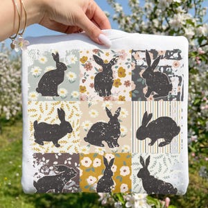 May include: White sweatshirt featuring a patchwork design of black rabbit silhouettes on various floral and patterned backgrounds. The design includes daisies, leaves, and striped patterns, creating a whimsical and artistic aesthetic.