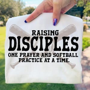 May include: White t-shirt with the text "RAISING DISCIPLES ONE PRAYER AND SOFTBALL PRACTICE AT A TIME." The design includes crossed baseball bats and a baseball. The text is in black, with the bats and baseball in a light gray.