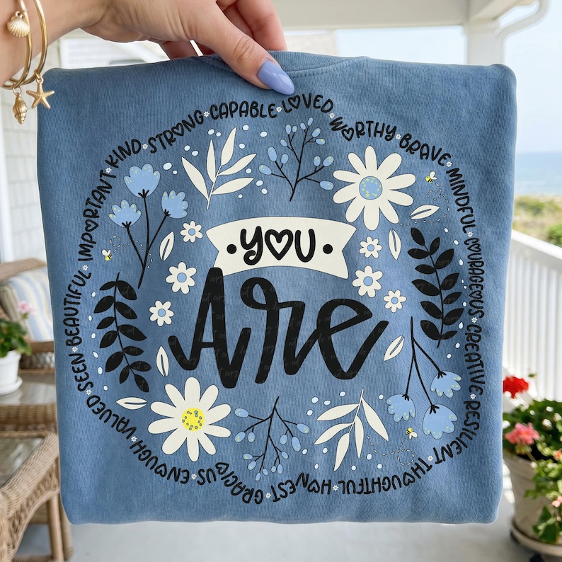 May include: A blue sweatshirt with the words "You Are" in a decorative font. The design includes a floral wreath with daisies, leaves, and inspirational words like "brave" and "worthy" around the edges.