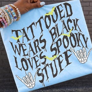 May include: Pale blue t-shirt featuring the words "TATTOOED WEARS BLACK SPOOKY LOVES STUFF" in black, with yellow bat graphics and skeleton hand gestures. The shirt is held up by a person.