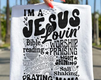 Peut inclure: Sweat-shirt blanc avec l'inscription noire "I'M A JESUS Lovin' MAMA". Le motif comprend les mots "Bible reading", "Worship", "Praising", "Kingdom Seeking" et "Praying". Le sweat-shirt est tenu par une main avec du vernis à ongles rouge.