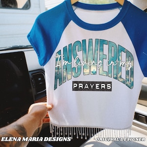 May include: White crop top with blue sleeves and neckline, featuring the text "ANSWERED I'm living in my PRAYERS" in a stylized font. The bottom hem is adorned with dangling silver beads. The words "ELENA MARIA DESIGNS" and "ORIGINAL DESIGNER" are also visible.