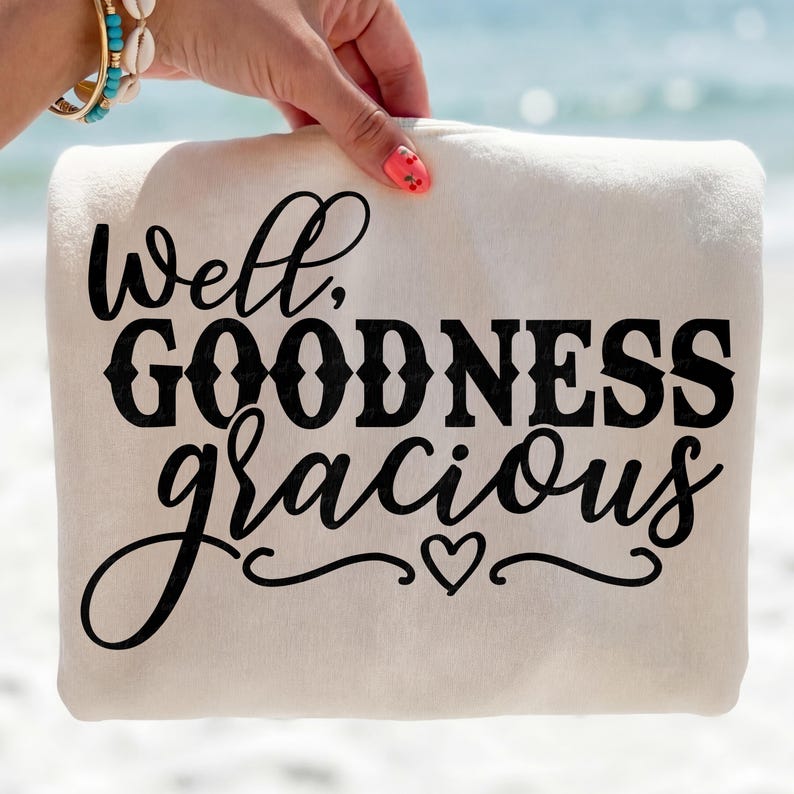 May include: A cream-colored beach towel with the phrase "Well, GOODNESS gracious" in black script. A small heart is at the bottom of the text. The towel is held by a hand with a pink manicure, with a turquoise and gold bracelet.