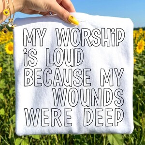 May include: White t-shirt with the text "MY WORSHIP IS LOUD BECAUSE MY WOUNDS WERE DEEP" in black block letters. The shirt is held up against a background of sunflowers and a blue sky.