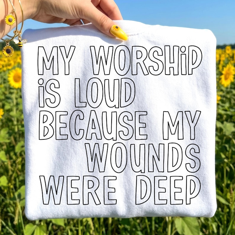 May include: White t-shirt with the text "MY WORSHIP IS LOUD BECAUSE MY WOUNDS WERE DEEP" in black block letters. The shirt is held up against a background of sunflowers and a blue sky.