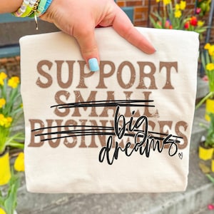 May include: Cream-colored t-shirt with brown text that reads "SUPPORT SMALL BUSINESSES" with a line through "SMALL BUSINESSES" and the words "big dreams" in black script. A small heart is next to "dreams".