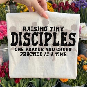 May include: A white t-shirt with the text "RAISING TINY DISCIPLES" in large, bold, black letters. Below, smaller text reads "ONE PRAYER AND CHEER PRACTICE AT A TIME." The shirt is held up against a backdrop of colorful flowers.