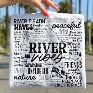 May include: White t-shirt with black text that reads "River Vibes." The design includes words like "adventure," "peaceful," "kayaking," and "sunshine." The shirt has a casual, outdoorsy theme.