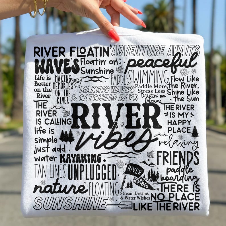 Puede incluir: Camiseta blanca con texto negro "River Vibes". El dise&ntilde;o incluye palabras como "aventura", "pac&iacute;fico", "kayak" y "sol". La camiseta tiene un tema informal y al aire libre.