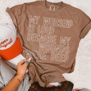 May include: A brown Comfort Colors t-shirt with the text "MY WORSHIP IS LOUD BECAUSE MY WOUNDS WERE DEEP" in white. A white and orange baseball cap is visible on the left.