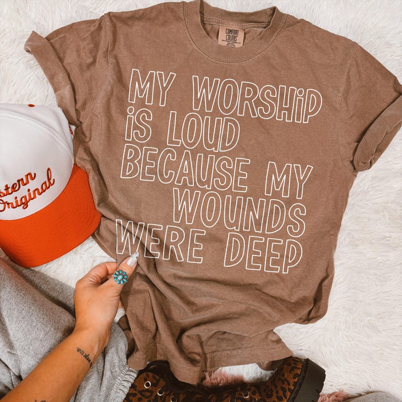 May include: A brown Comfort Colors t-shirt with the text "MY WORSHIP IS LOUD BECAUSE MY WOUNDS WERE DEEP" in white. A white and orange baseball cap is visible on the left.
