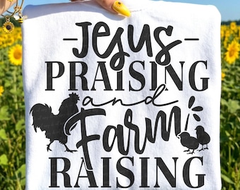 May include: White t-shirt with the text "Jesus Praising and Farm Raising" in black. A black rooster and two chicks are also featured on the shirt. The shirt is held up in front of a field of sunflowers.