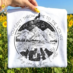 May include: White sweatshirt with a black and white graphic of a wolf howling on a mountain, surrounded by a circle with the text "YOU CAN'T THROW ME TO THE WOLVES, THEY COME WHEN I CALL". The design includes mountains, a lake, and other wolves.