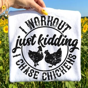 May include: White t-shirt with a black graphic design. The design features the text "I WORKOUT just kidding I CHASE CHICKENS" in a circular arrangement around two black chicken silhouettes. The shirt is held up against a background of sunflowers.